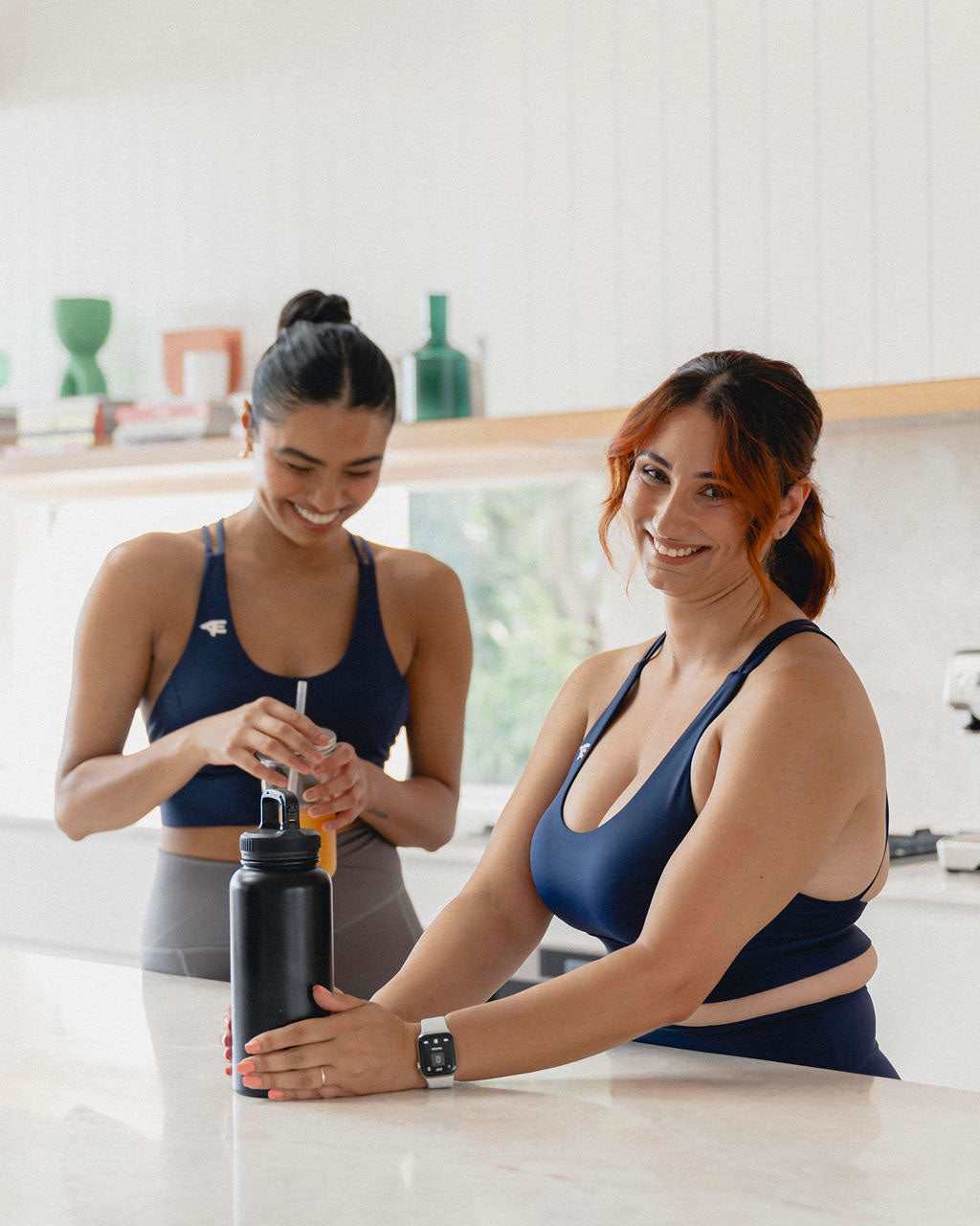 Two women wearing matching navy blue sports bras stand in a bright, modern kitchen. One smiles while holding a water bottle, while the other prepares a drink, both exuding a relaxed and friendly vibe, ready for their fitness routine. Horizontal view