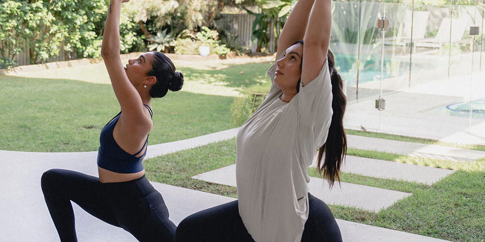 Two women in a yoga lunge pose on mats outdoors, with arms raised, surrounded by greenery and a modern backyard.