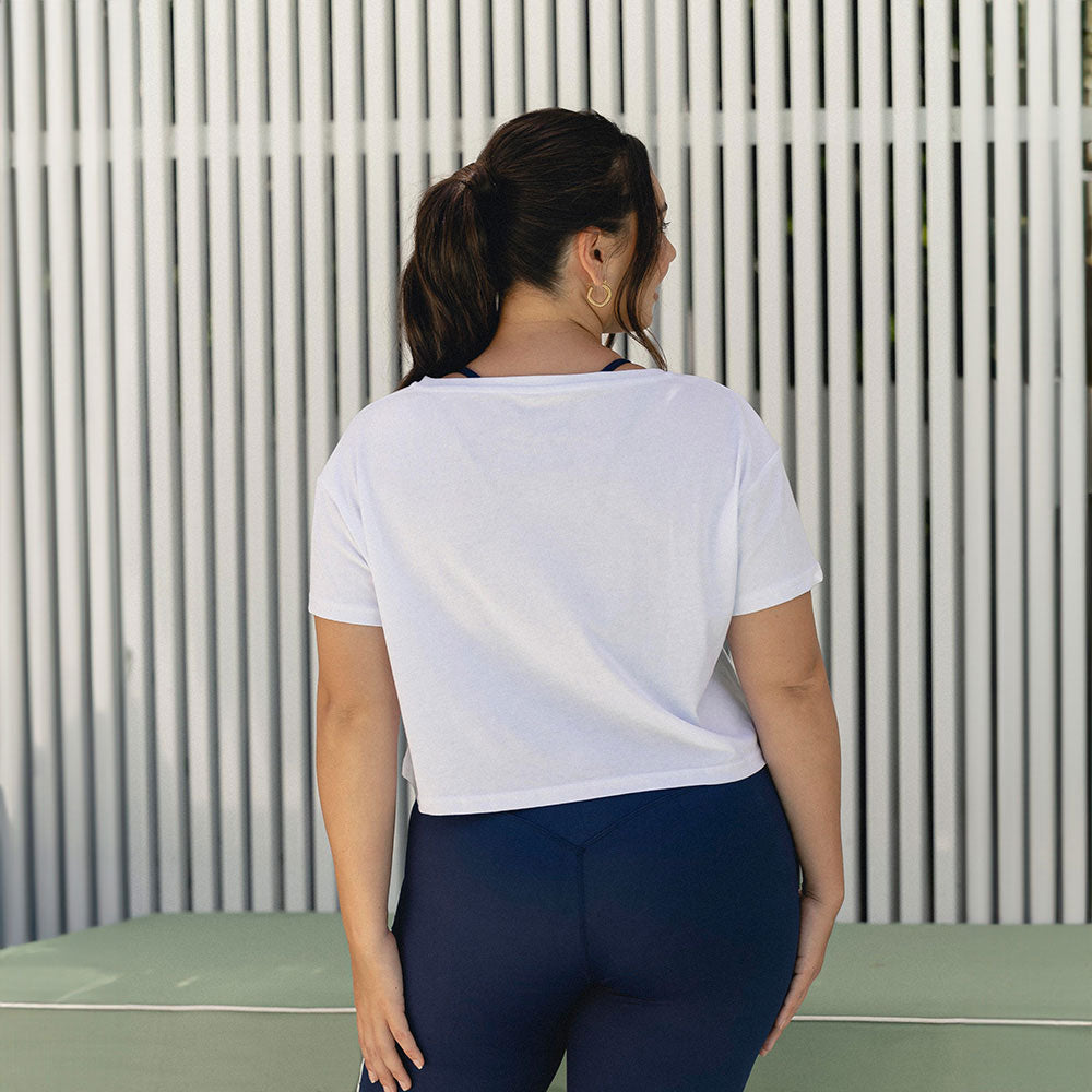 A woman wearing a white cropped fitness top, viewed from the back, standing confidently in front of a minimalist backdrop.