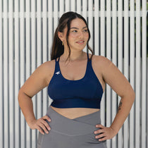 A woman confidently posing in a navy sports bra and grey leggings, radiating strength and body positivity in a minimalist backdrop.