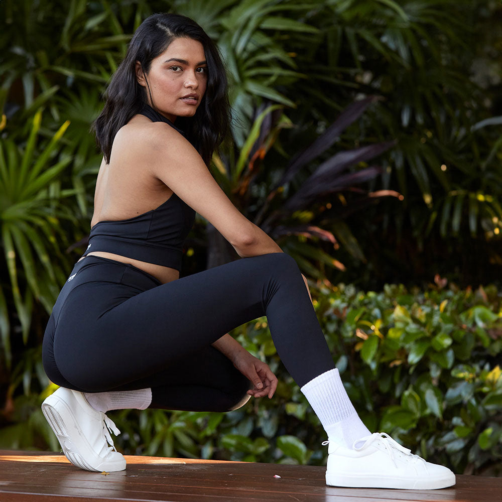 A woman squatting in a stylish black activewear set, consisting of a halter top and leggings, looking confidently at the camera amidst a natural backdrop of greenery.