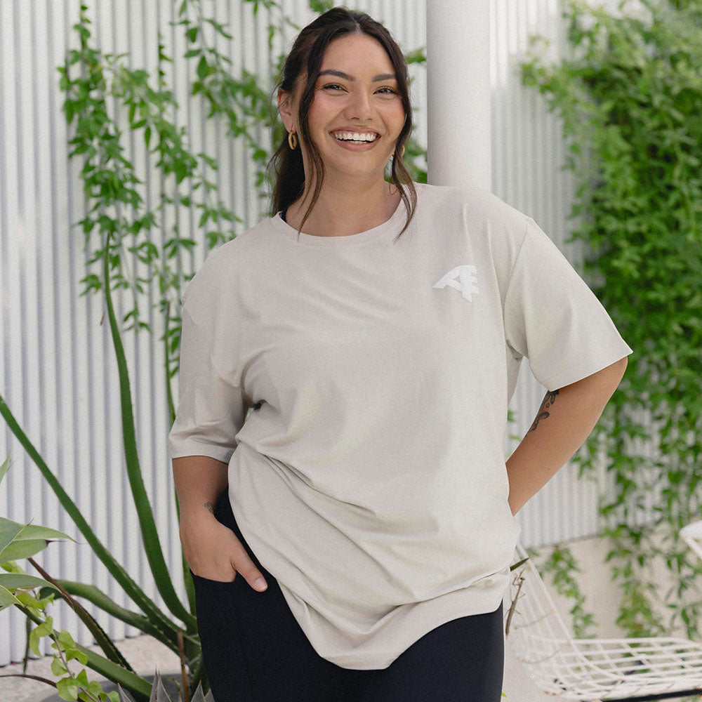 A smiling woman wearing an oversized sandy coloured Active Elite t-shirt, posed in a vibrant outdoor setting, surrounded by plants. She looks comfortable and at ease, showcasing a casual and sporty style.