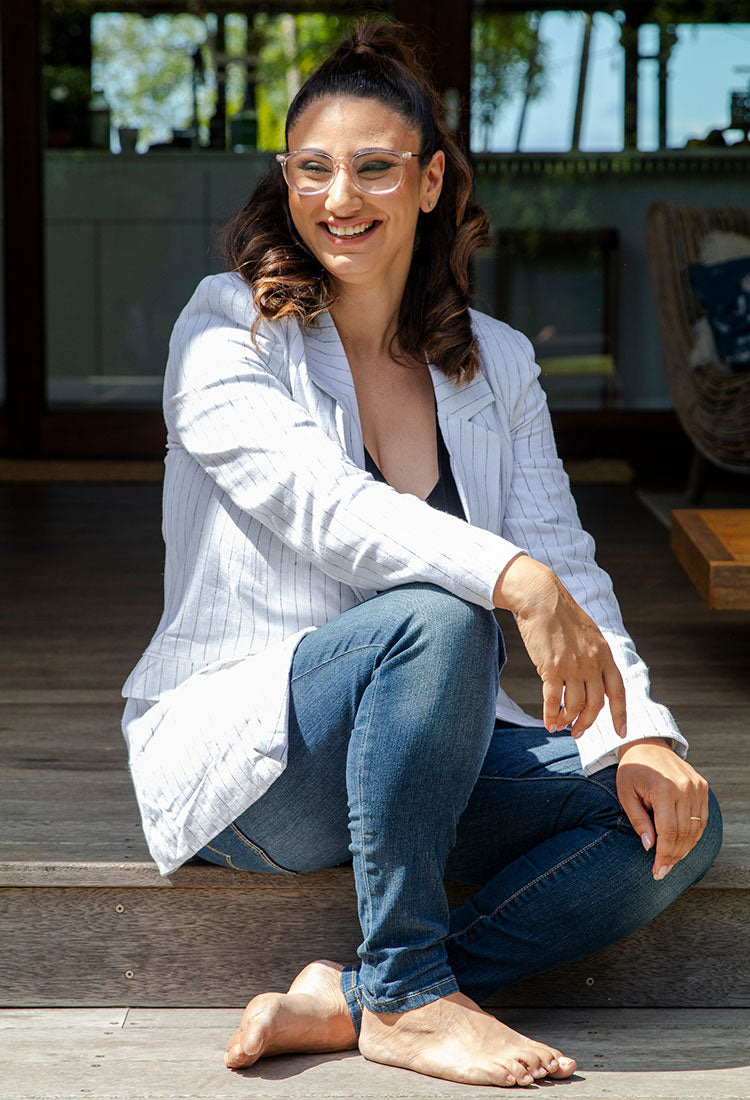 Professional woman in white pinstriped blazer and glasses sitting casually on wooden deck, wearing blue jeans, natural sunlit business portrait