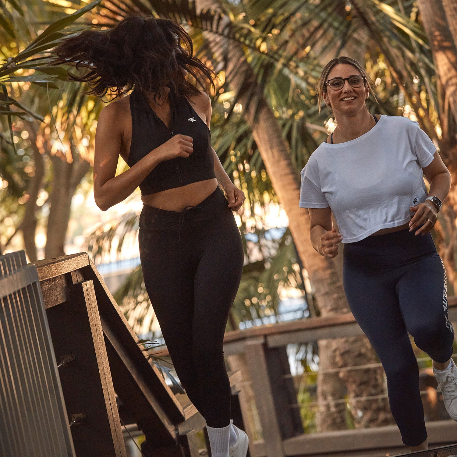 Two women jogging outdoors in a tropical setting. One wears a black halter top and leggings, while the other sports a white cropped t-shirt and blue leggings, both enjoying an energetic run.