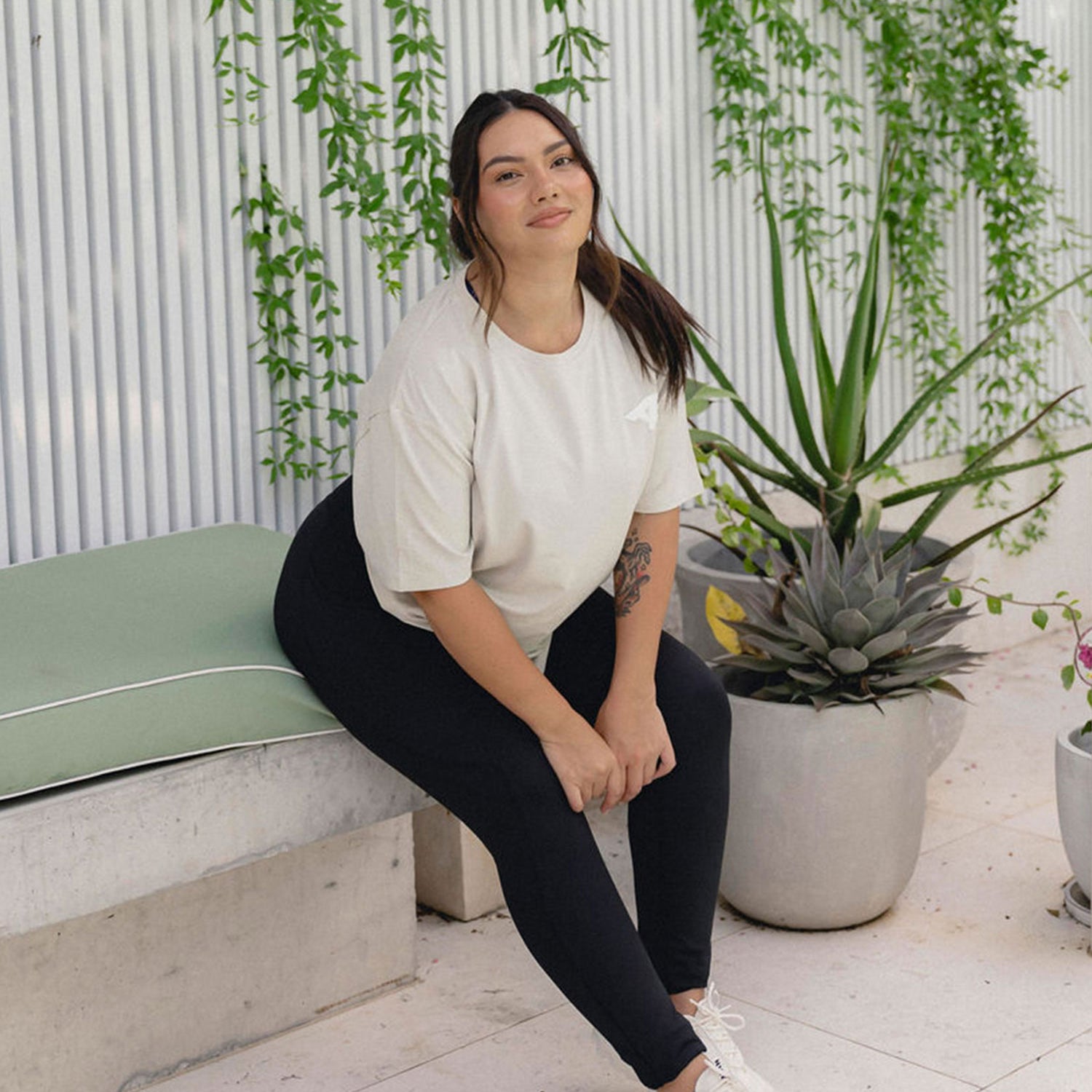 A young woman sits on a bench in a relaxed outdoor setting with greenery. She's wearing a light oversized t-shirt, black leggings, and white sneakers, smiling softly with plants in the background.