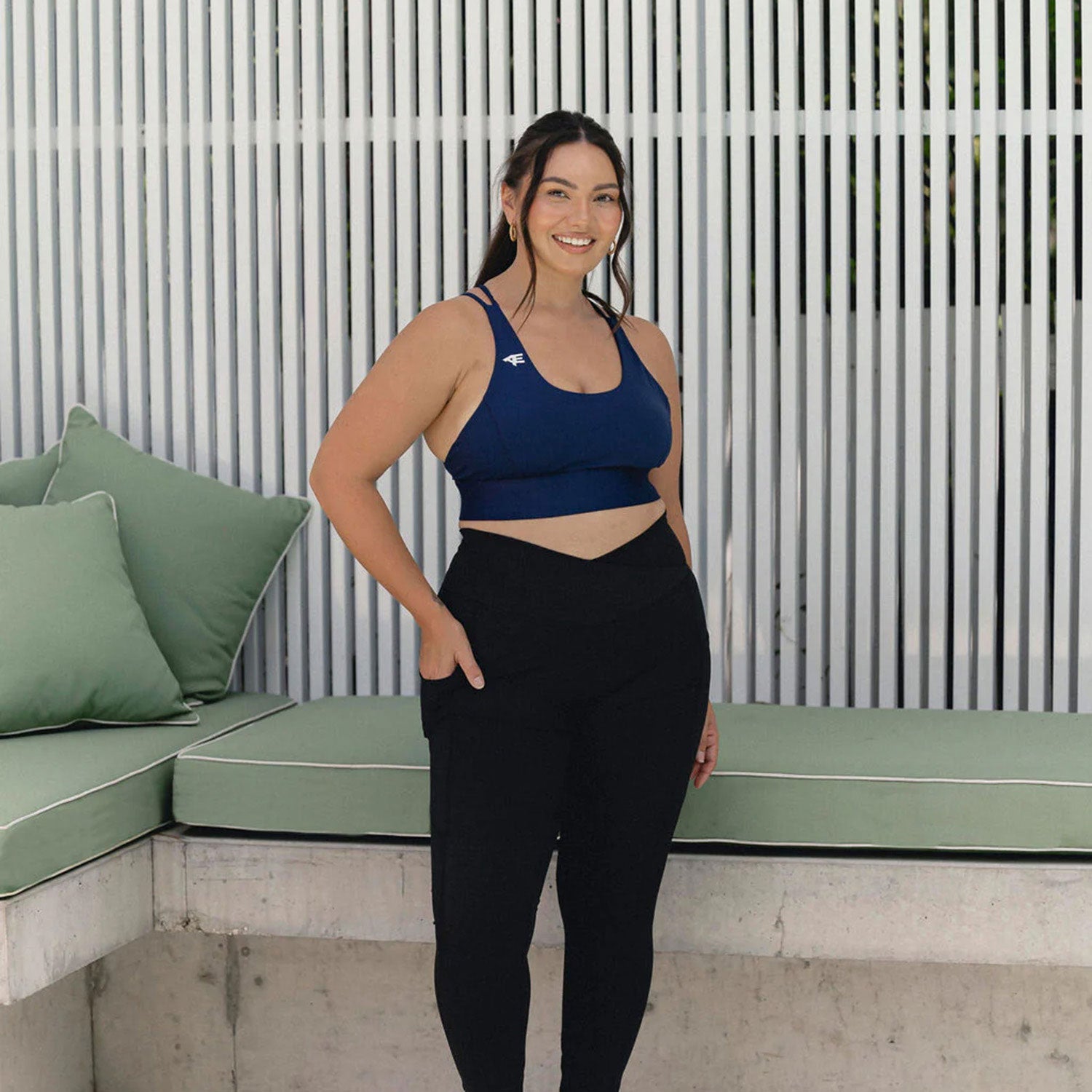 Woman standing confidently in activewear, featuring a navy blue sports bra and black leggings, in front of a stylish outdoor seating area.