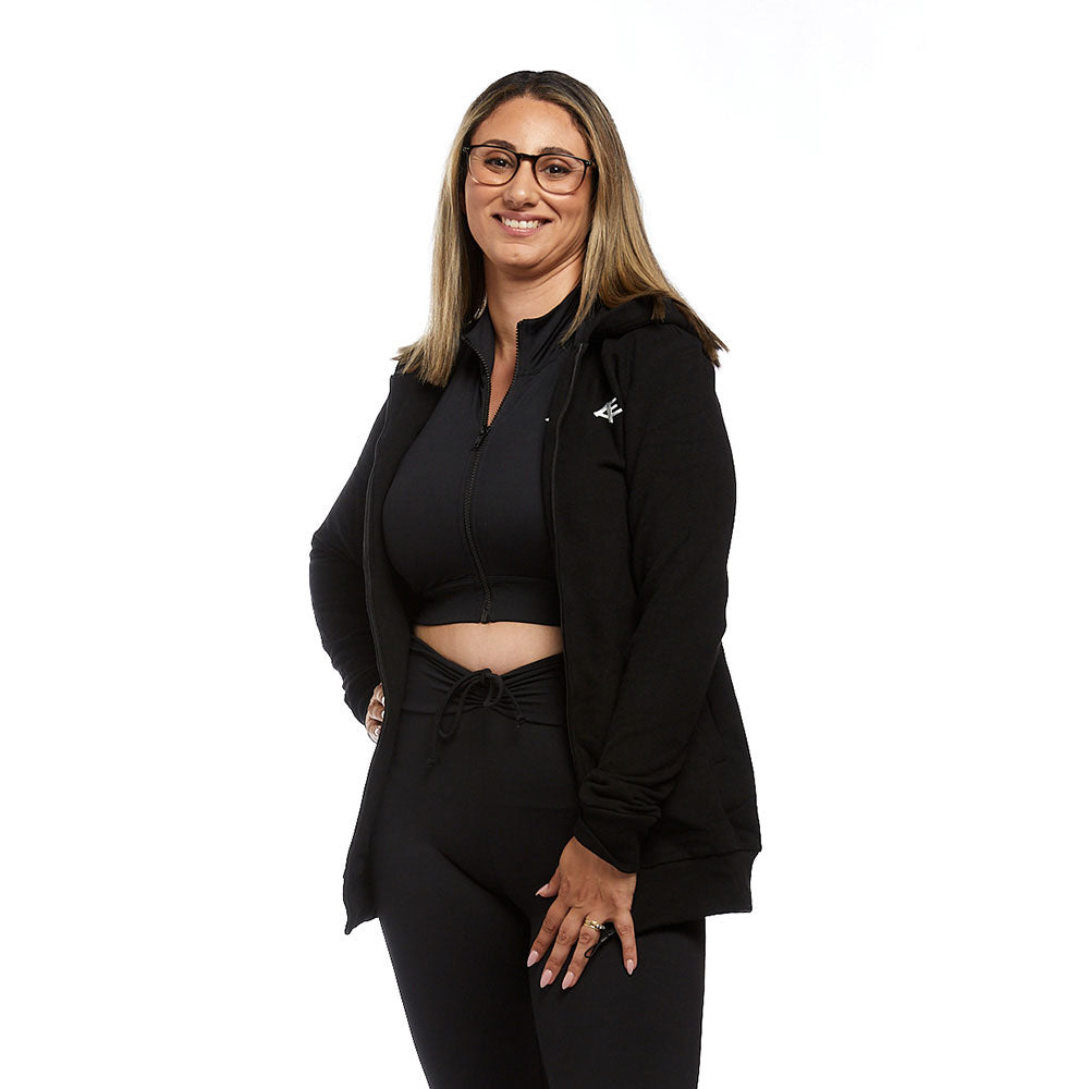 A woman smiling while wearing a black zip-up Active Elite hoodie, open to reveal a crop top underneath. She pairs the hoodie with black leggings, showing off a stylish and comfortable athleisure outfit.